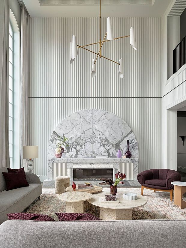 A refined, high-ceiling living room with dramatic floor-to-ceiling fluted wall panelling, a large arched marble fireplace surround, and tall windows with sheer curtains. The room is furnished with a large grey sofa, burgundy upholstered armchairs and cushions, sculptural pale stone coffee tables, and a contemporary brass-and-white pendant light. A mezzanine or upper-level balcony with dark balustrading is visible at right.