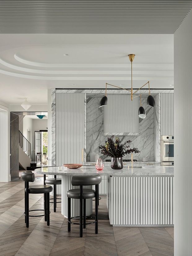 A sophisticated contemporary kitchen with a large marble island, vertically fluted cabinetry, marble splashback, integrated oven, and sculptural brass-and-black pendant lighting. Black bar stools surround the island, while a chevron timber floor and curved layered ceiling details add texture. The open-plan space connects visually to a stairway and adjacent living areas.