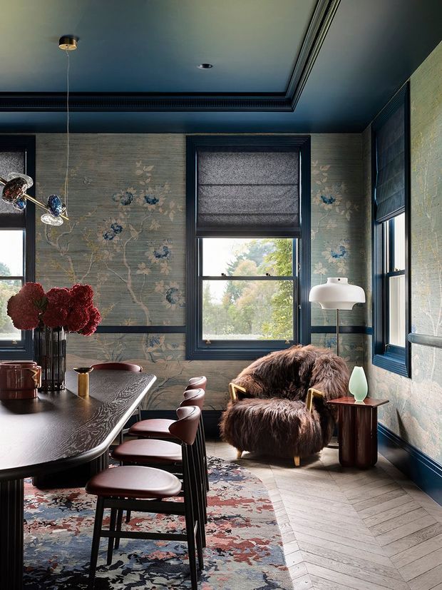 A richly layered dining room with dark blue painted trim, a coffered blue ceiling, patterned floral wallcovering, and multiple tall windows with roller blinds. A dark sculptural dining table is paired with upholstered dining chairs, while a shaggy brown lounge chair and side table occupy the window corner. A patterned rug, floral arrangement, pendant light, and floor lamp add texture and colour.