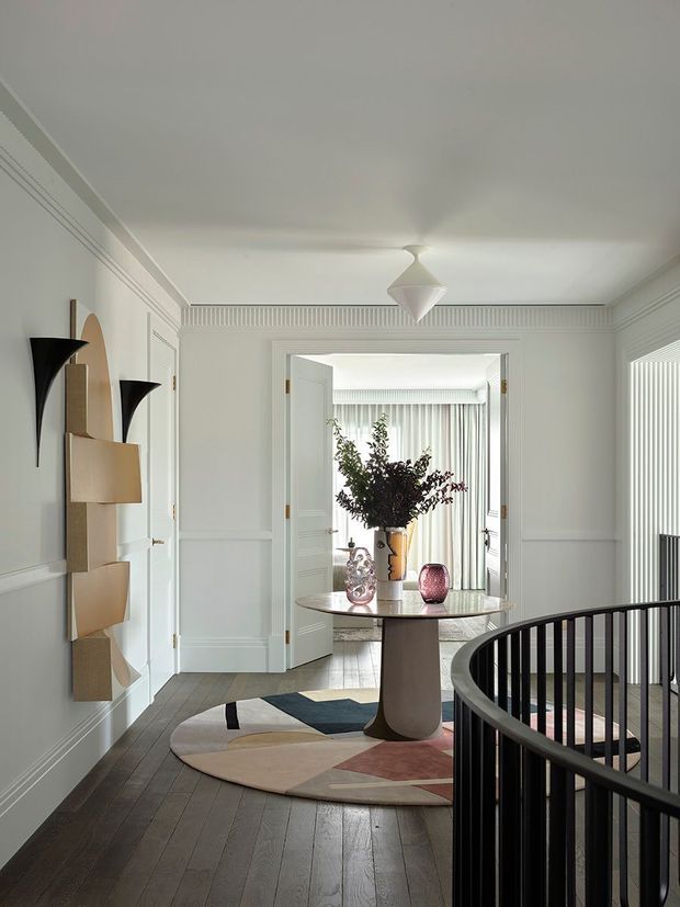 Refined bright interior landing or hallway with white decorative wall panelling, high ceiling, dark timber flooring, a sculptural black stair balustrade, and a round console table decorated with glass vessels and foliage. A geometric area rug adds muted blue, beige, pink and charcoal accents. The space opens into a softly furnished room beyond and features contemporary sculptural wall lighting and a faceted ceiling pendant.