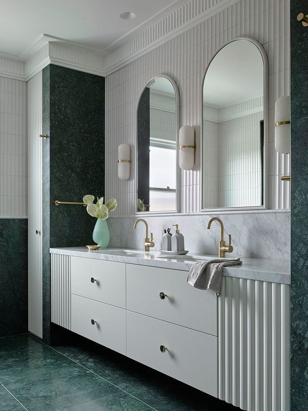 Elegant bathroom vanity with a long double-sink cabinet, white marble-look benchtop and splashback, arched mirrors, ribbed white wall panelling and cabinetry, brass tapware and sconces, and dark green stone-look wall and floor surfaces. The refined scheme combines classic detailing with contemporary bathroom fittings.