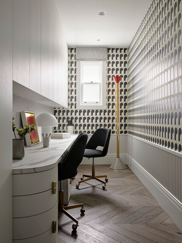 A narrow, stylish home office with a built-in curved white desk and cabinetry along the left wall, two black upholstered swivel chairs, a marble-look desktop, and a tall floor lamp. Bold repeating black-and-white patterned wallpaper covers the right and rear walls, while herringbone timber flooring, white wall panelling, and a small window create a refined, graphic interior.
