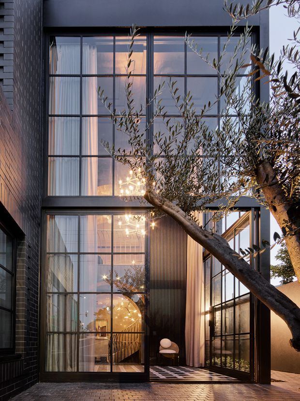 A dusk exterior view of a contemporary multi-storey residence with a dark brick facade, extensive black-framed floor-to-ceiling glazing, and a tall glazed entry or living space. Warm interior lighting reveals curtains, a sculptural staircase, pendant lighting, and a small seating area. An olive tree crosses the foreground, and the ground-level entry is paved in brick with a patterned threshold.