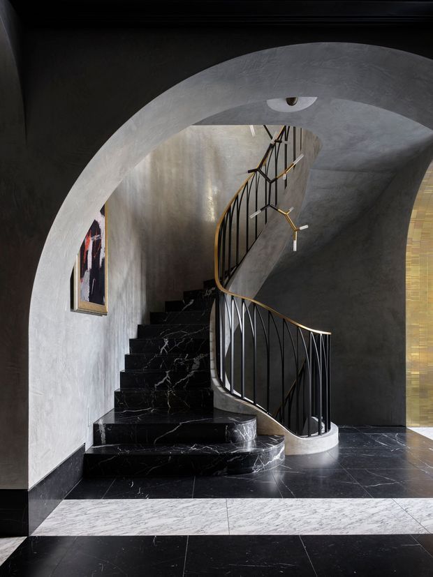 Dramatic interior stairway framed by a large plastered arch, featuring sweeping curved black marble stairs and flooring, dark grey textured walls, and an elegant black metal balustrade with brass-toned handrail details. A sculptural branching pendant light hangs beside the stair, while a narrow vertical window and artwork add small accents.