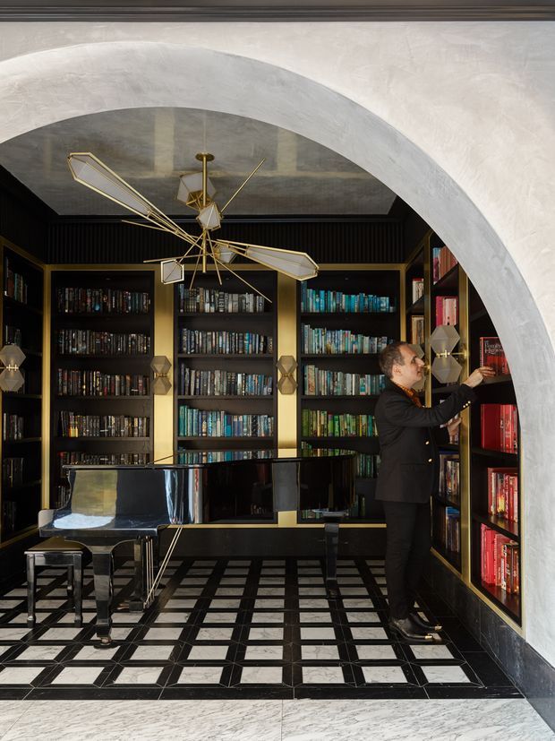 A dramatic, richly detailed library or music room viewed through a large white plaster arch. Floor-to-ceiling black built-in bookshelves with brass-gold framing line the room, filled with colourful books. A glossy black grand piano and bench sit on a geometric black-and-white tiled floor. The dark ceiling has ribbed detailing and a sculptural brass geometric pendant light hangs centrally. A person is browsing the shelving.