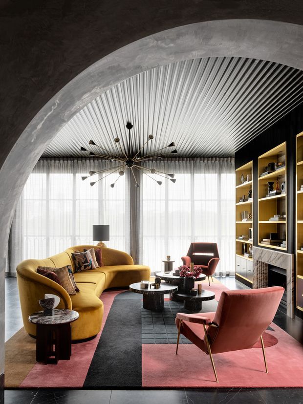 A sophisticated living room framed by a large curved concrete arch, featuring a dramatic ribbed metallic ceiling, full-height sheer-curtained glazing, a mustard-yellow curved sofa, rose-pink lounge chairs and rug, sculptural black coffee tables, and a tall built-in shelving and media unit in warm yellow and dark tones. A striking mid-century-inspired starburst chandelier forms the central focal point.