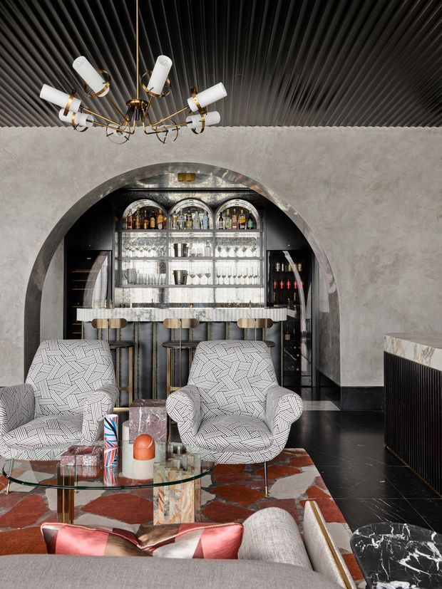 A richly styled lounge and home bar with a dramatic arched opening, mirrored and glass-backed bottle display, bar counter with stools, patterned armchairs, a glass coffee table, and layered rugs. The room features black ribbed ceilings, pale textured plaster walls, dark flooring, brass lighting and details, and high-contrast geometric furnishings.