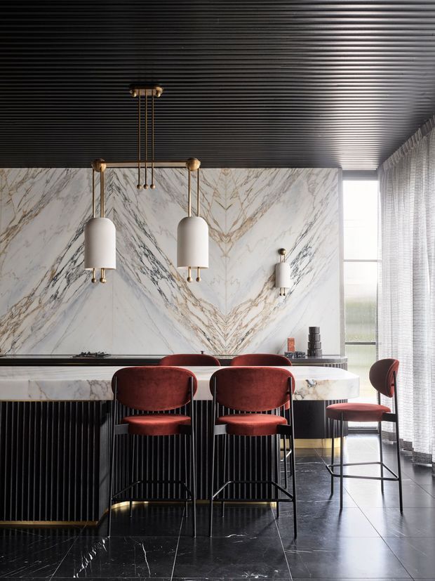 A dramatic contemporary kitchen with a long marble island and matching veined marble splashback, black ribbed cabinetry, dark tiled flooring, and deep burgundy upholstered bar stools. Sculptural brass-and-white pendant lights hang over the island, while sheer curtains and tall glazing bring in natural light. The room has a refined, high-contrast, luxurious atmosphere.
