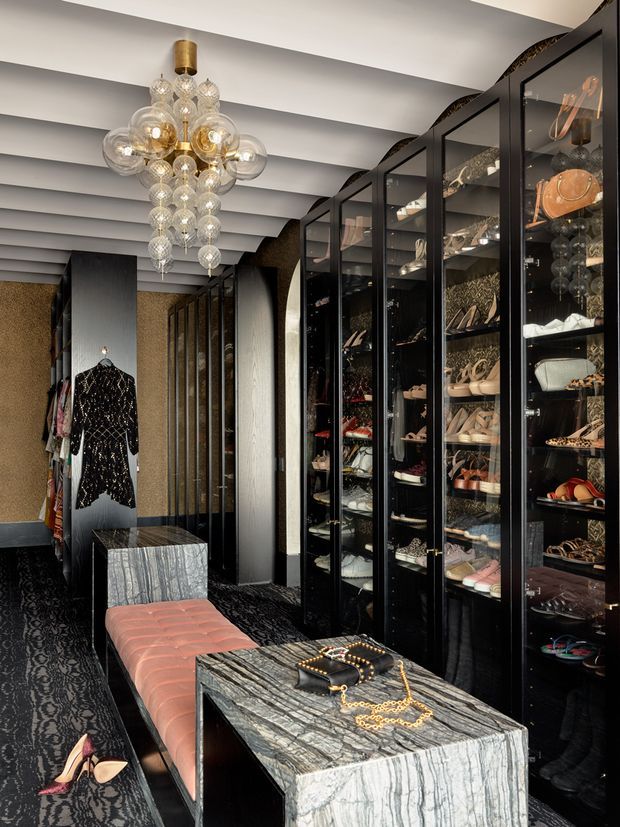A glamorous walk-in wardrobe or dressing room with floor-to-ceiling black-framed glass cabinetry displaying shoes, handbags and accessories. The space features dark patterned carpet, dramatic black and metallic finishes, veined stone display tables, a blush upholstered bench, a sculptural cluster pendant light, and a hanging black embellished garment.
