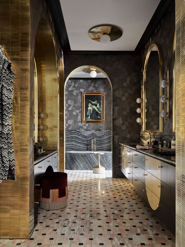 A dramatic, richly detailed bathroom with a long double-vanity layout, dark geometric wall finish, brass-framed arched mirrors, marble vanity surfaces, and a freestanding bathtub visible through the central arch. The space features patterned terrazzo-like floor tiles, warm metallic accents, globe lighting, and layered decorative styling.