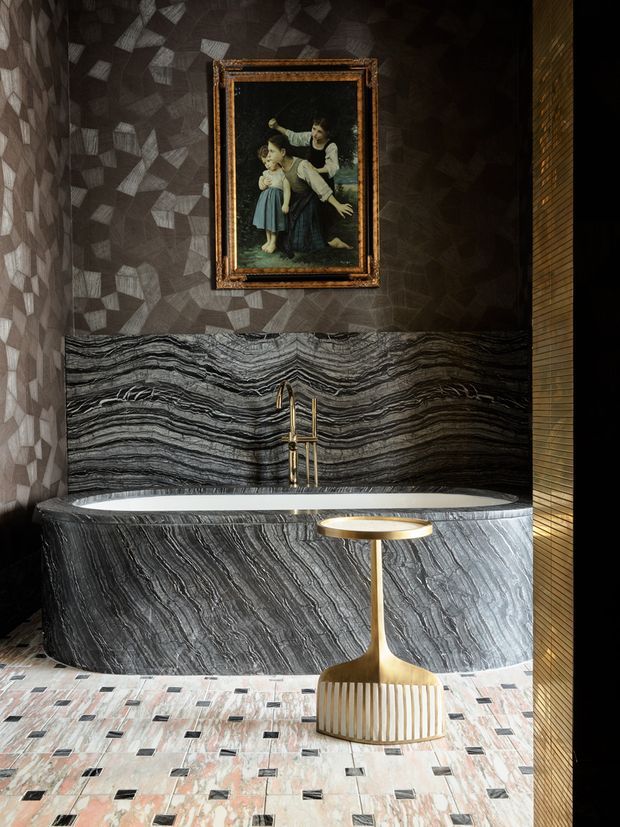 A dramatic, luxurious bathroom centered on a freestanding oval bathtub clad in richly veined dark grey stone. The bath sits against a matching stone feature wall with a sculptural brass tap, while a small brass side table and chair occupy the foreground. Dark geometric wall covering, framed artwork, warm metallic detailing, and a patterned stone-tile floor create an opulent, art-deco-inspired interior.