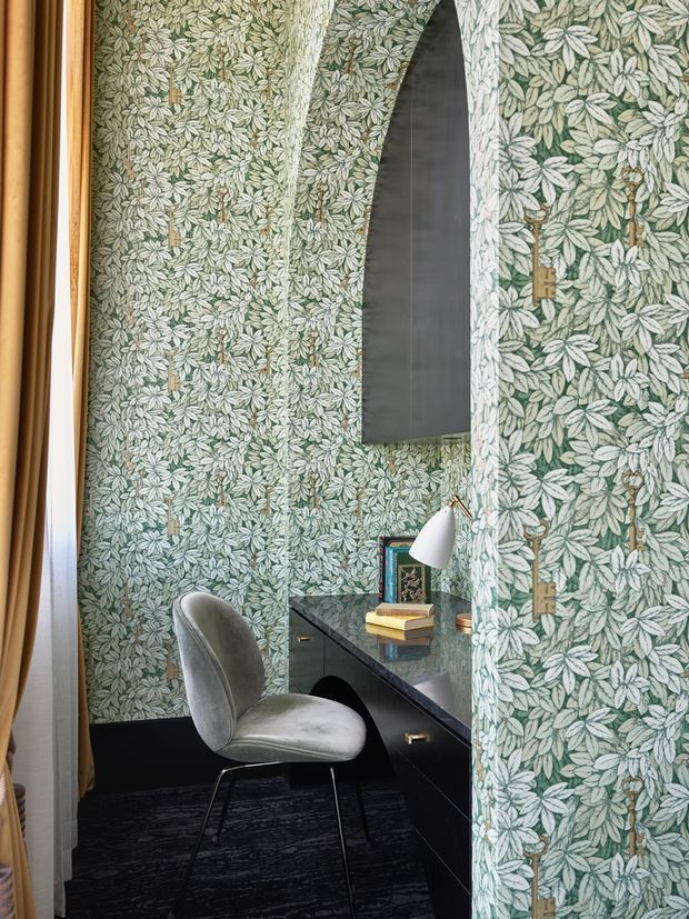 A compact, enclosed home office nook with floor-to-ceiling botanical wallpaper in muted green and cream, a dark built-in desk with glossy stone-like surface and drawers, and a curved grey upholstered chair. A wall-mounted sculptural black feature light or cabinet rises above the desk, while a small white desk lamp, books and framed artwork add functional detail. Curtains frame the space on the left, and dark carpet covers the floor. The room has a richly patterned, refined and intimate character.