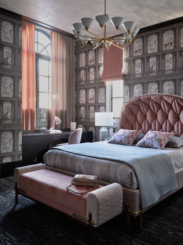 A richly decorated bedroom with a large upholstered bed, dramatically tufted blush-pink headboard, grey-blue bedding, and a matching upholstered bench at the foot. The room features ornate dark grey patterned wallcovering with classical architectural motifs, tall arched windows dressed in sheer peach curtains, a dark desk and chair, sculptural bedside lighting, and a large contemporary brass-and-white chandelier. The overall design is glamorous, theatrical, and highly detailed.