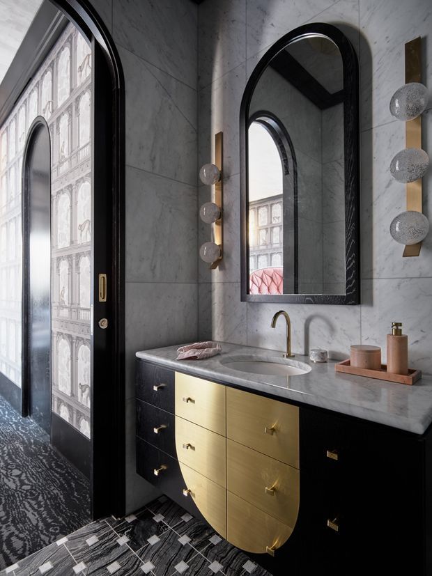 A dramatic, compact bathroom or powder room with a marble-clad wall and countertop, a curved black-framed arched mirror, arched black door, brass faucet, and a striking black-and-gold built-in vanity. Decorative globe wall lights and patterned dark flooring add an artful, luxurious character. Architectural detailing is also visible in the adjacent reflected and glazed surfaces.