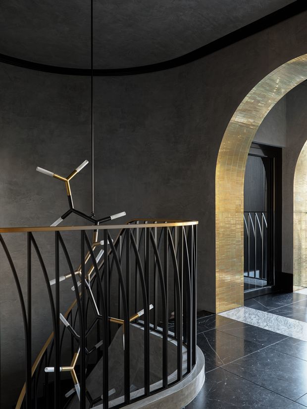 A dramatic, refined stairway or landing with a curved black metal balustrade, dark textured plaster walls and dark tiled flooring. A sculptural branching pendant light hangs over the stair opening, while a large rounded arch is lined with shimmering gold-toned mosaic tiles and frames a dark doorway.