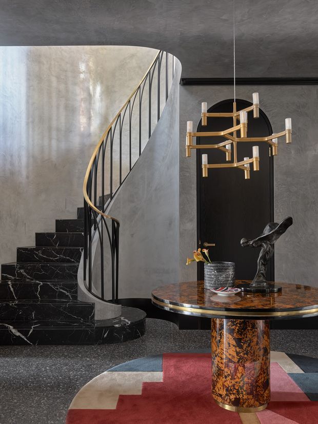 A dramatic, high-end entry foyer featuring a sweeping curved staircase with black marble treads, a slim black metal balustrade and brass handrail. Dark textured plaster walls and ceiling surround a black arched door. A sculptural brass chandelier hangs above a round table with a richly patterned stone or marble top and ornate cylindrical pedestal. A geometric red, blue and cream rug and sculptural decor complete the moody, artful space.