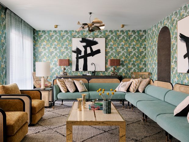 A richly layered living room featuring a large curved green sectional sofa, mustard velvet armchairs, patterned floral wallpaper, a geometric rug, and a rectangular timber coffee table. A painted abstract artwork, woven and timber details, table lamps, and a sculptural ceiling fan create an eclectic, colourful, sophisticated interior.