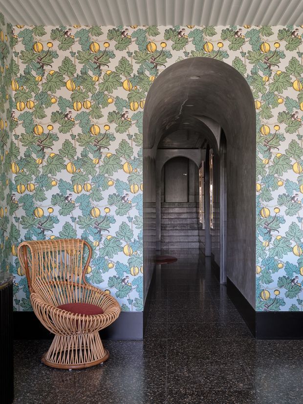 A moody interior hallway featuring bold botanical wallpaper with yellow fruit and blue-green foliage, a dark arched doorway leading to a stair hall, dark polished terrazzo flooring, black wall skirting, and a sculptural woven rattan chair with a burgundy cushion. The ceiling has narrow ribbed panels, creating a dramatic, richly patterned transitional space.