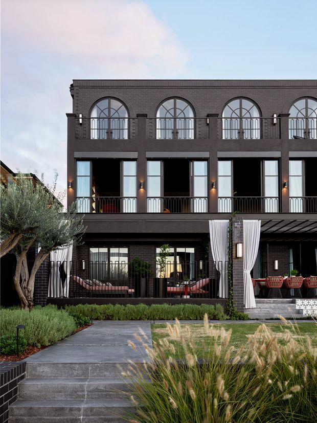 Front exterior view of a multi-storey dark brick townhouse or apartment-style residence, featuring repeated arched upper-level windows, tall rectangular glazing, metal balcony railings, covered outdoor entertaining areas with curtains, and a landscaped garden with a paved path and steps.