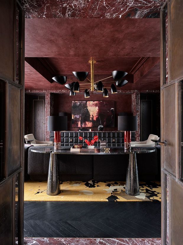 A dramatic, richly layered interior viewed through dark timber-framed doors. The room features deep burgundy marble and plaster surfaces, black herringbone flooring, a large dark dining or console table with sculptural metallic legs, black upholstered seating, statement artwork, red floral accents, and a sculptural black-and-brass pendant light. The overall composition is luxurious, moody, and Art Deco-inspired.