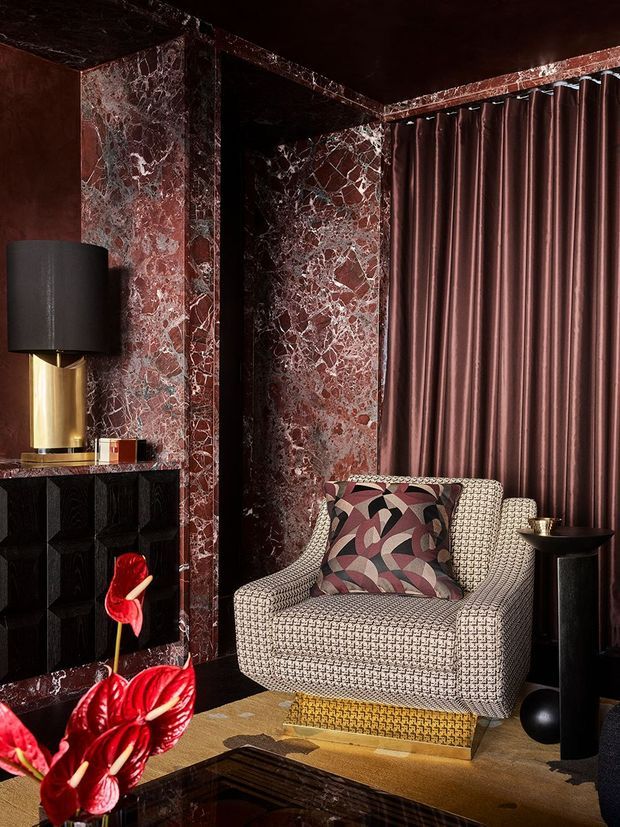 A richly styled, moody interior corner featuring a sculptural upholstered armchair with a patterned cushion, set against dramatic burgundy marble wall panels and matching velvet curtains. A black geometric built-in cabinet, brass-and-black table lamp, dark pedestal table, reflective coffee table, and red anthurium flowers create a luxurious Art Deco-inspired atmosphere.