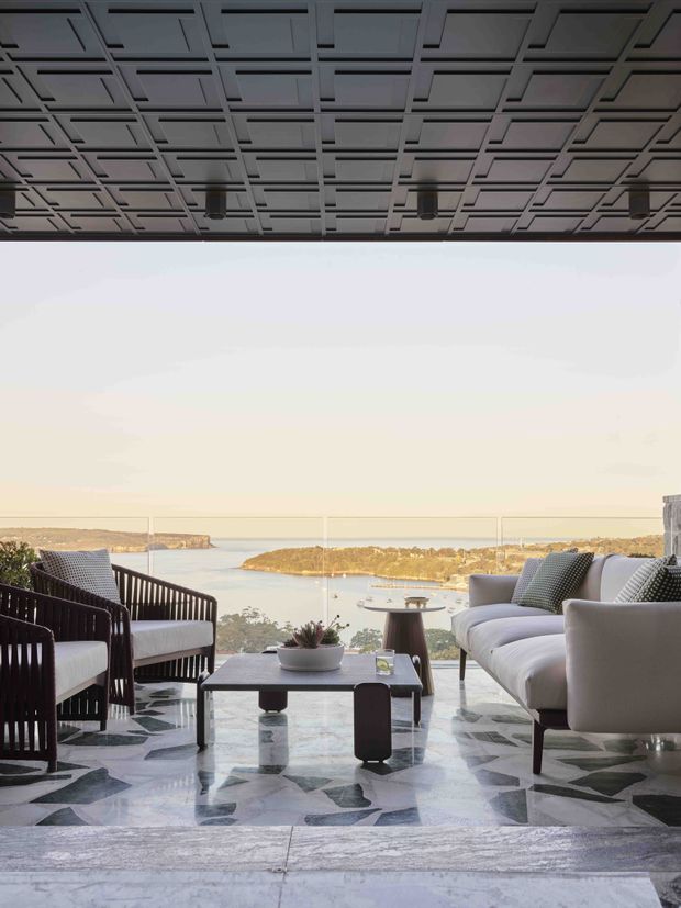 A sophisticated open-air or glazed living lounge overlooking a coastal bay, furnished with a large pale sofa, dark woven armchairs, a low coffee table, and a small side table. The space features a dramatic geometric coffered ceiling, full-width glass balustrade or glazing, and a striking irregular stone-patterned floor in grey, white, and dark tones. The elevated view includes water and rocky coastal landforms in the distance.