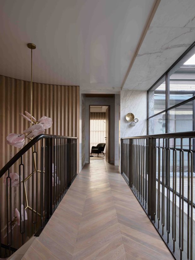 A refined upper-level hallway or landing with a warm herringbone timber floor, vertical timber wall panelling, dark metal balustrades, and a large marble-clad wall beside expansive black-framed glazing. A doorway leads to a small seating area, while sculptural wall lights and delicate floral decor add a polished, contemporary character.