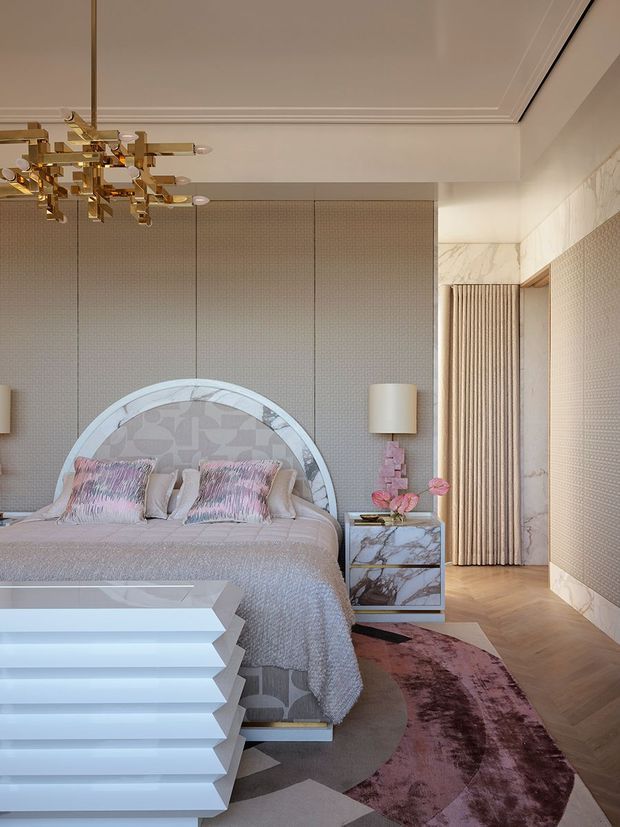 A luxurious, highly styled bedroom with a large upholstered bed framed by a decorative circular arch, patterned headboard, blush and cream bedding, and sculptural geometric bedside furniture. The room features marble-look surfaces, textured taupe wall panels, herringbone timber flooring, a plush abstract mauve rug, pleated drapery, ornate white ceiling mouldings, and a striking brass-and-glass chandelier. The overall aesthetic is glamorous, refined, and strongly Art Deco-inspired.