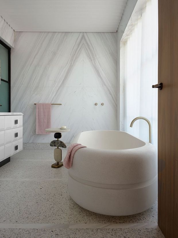 Bright contemporary bathroom featuring a rounded freestanding bathtub, pale veined marble-look wall panels, terrazzo flooring, white vanity cabinetry, a brushed brass-toned curved tap, and a large sheer-covered window. A timber door and soft pink towel add warmth and contrast.