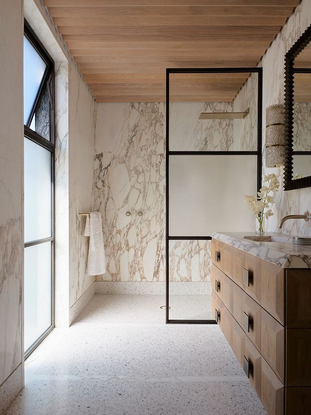 A refined contemporary bathroom with a walk-in shower enclosed by a black-framed glass and textured privacy screen. The space features veined marble-look wall surfaces, a pale terrazzo floor, warm timber-look ceiling panels, and a timber vanity with a matching veined stone benchtop. A large window provides abundant natural light, while a sculptural dark-framed mirror and brushed metal tapware add contrast.