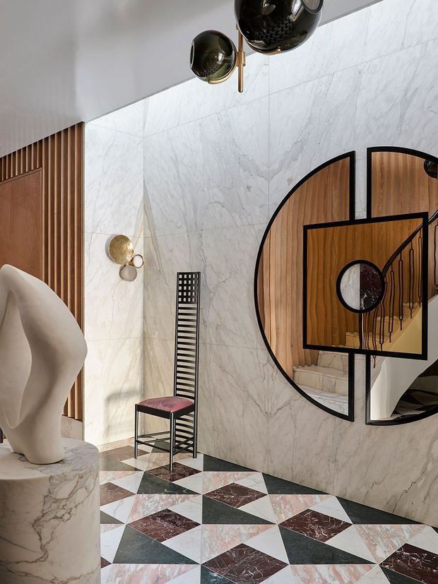 A dramatic, artfully detailed interior foyer or hallway featuring full-height veined marble wall cladding, geometric patterned marble flooring, warm vertical timber wall slats, and a striking black-framed circular and arched mirror reflecting a staircase. Sculptural lighting, a sculptural white pedestal object, and a slender decorative chair add an art-deco-inspired character.