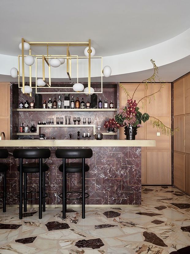 A sophisticated home bar with a long stone-topped counter, black bar stools, extensive bottle and glass storage, and a dramatic purple-brown marble bar front and backsplash. Brass geometric pendant lighting, warm timber cabinetry, a curved white ceiling, and patterned stone flooring create a luxurious, sculptural interior.