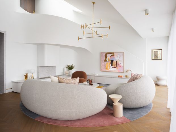 Bright, refined living room with sculptural curved boucle sofas arranged on a layered circular rug. The white interior features softly rounded built-in architectural forms, pale timber herringbone flooring, minimalist white cabinetry, a contemporary abstract artwork, and a delicate brass linear pendant light. High-level glazing or a skylight brings in abundant natural light, creating an airy contemporary setting.