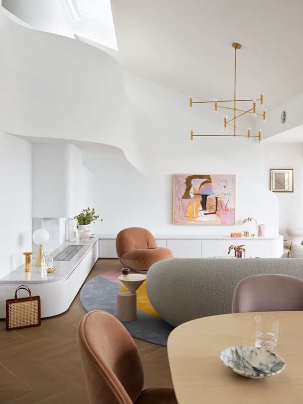 Bright, refined open-plan living and dining room with sculptural white architectural forms, a white built-in kitchen or bar area, warm herringbone timber flooring, curved contemporary seating, a rounded dining table, colourful abstract artwork, and a linear brass pendant chandelier. The room has abundant natural light from a skylight or clerestory glazing and a soft, sophisticated neutral palette with warm leather accents.
