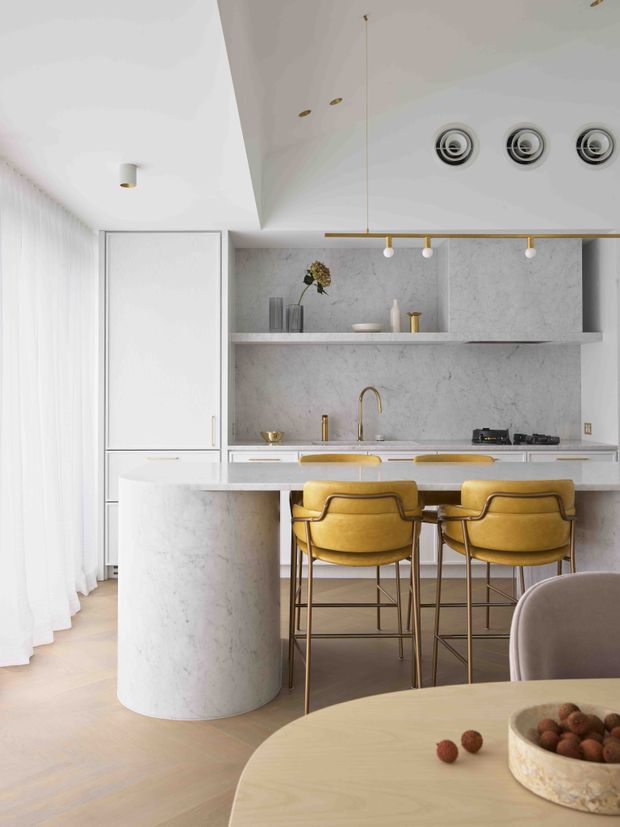 Bright contemporary kitchen with a curved marble island and integrated dining counter, white marble-look cabinetry and splashback, brass-toned hardware and fittings, and two mustard-yellow bar stools. Sheer curtains, pale timber flooring, recessed ceiling lights, and a linear pendant create an airy, refined interior.