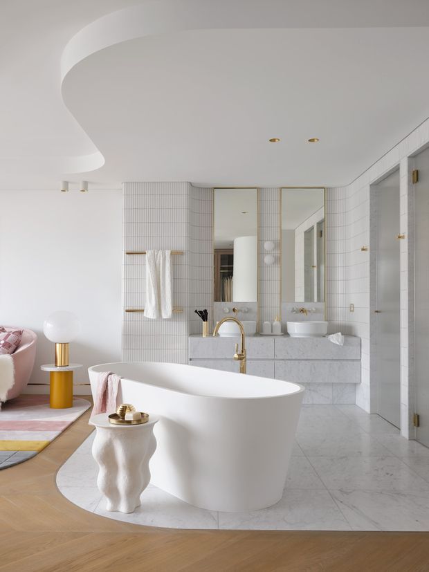 A bright, luxurious bathroom with a freestanding white bathtub in the foreground, double vanity with vessel sinks and tall mirrors, tiled walls, marble-look vanity cabinetry, and a walk-in shower. The room includes curved ceiling detailing, brass fixtures, and a small adjoining seating area with a pink sofa visible at left.