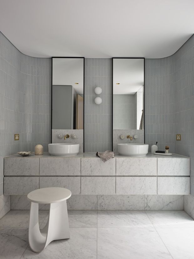 A refined contemporary bathroom with a double vanity, two vessel basins, tall black-framed mirrors, and wall-mounted brass fixtures. The room is finished in pale grey vertically ribbed wall tile and large-format white marble-look tiles across the vanity, cabinetry fronts, and floor. A sculptural white stool sits in the foreground, while recessed curved ceiling transitions soften the room's geometry.