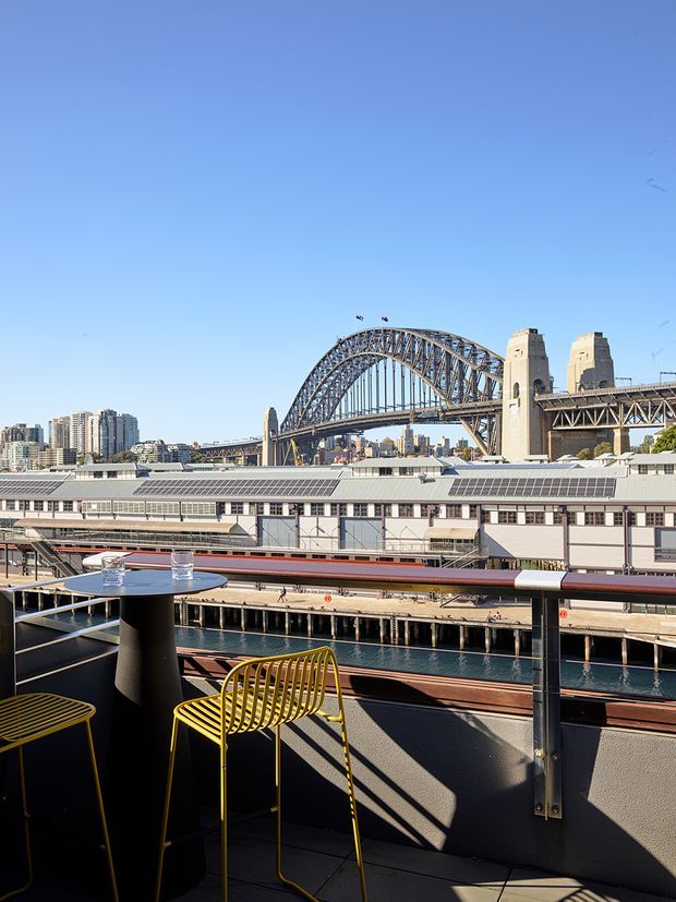A sunlit urban terrace or balcony overlooking a harbour, with a striking steel arch bridge and historic stone bridge towers in the background. The foreground includes a compact black outdoor table, yellow metal bar stools, dark balustrades, and a tiled or paved terrace surface. A waterfront industrial or transport building and city apartment towers are visible beyond.
