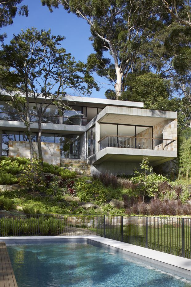 Exterior view of a contemporary multi-level residence set into a lush landscaped site, with projecting concrete balconies, extensive glazing, pale stone wall sections, and a swimming pool in the foreground. Mature trees and garden planting surround the house.