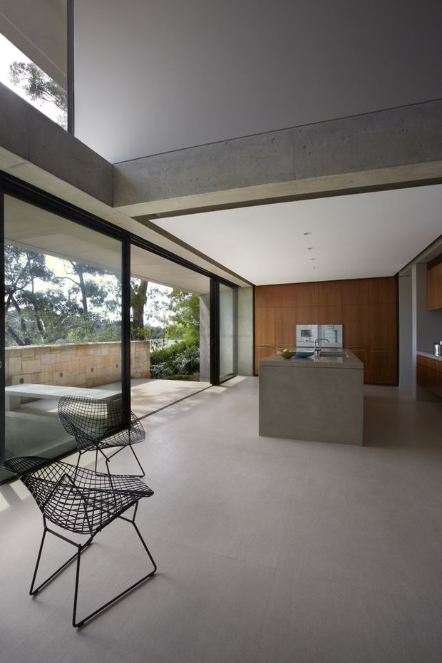 A refined open-plan kitchen and living area with a large concrete island, warm timber cabinetry, expansive black-framed glazing, and a strong indoor-outdoor connection to a covered terrace and landscaped garden. Exposed concrete beams and a minimalist ceiling structure define the architectural character, while wire-frame lounge chairs add a sculptural mid-century touch.