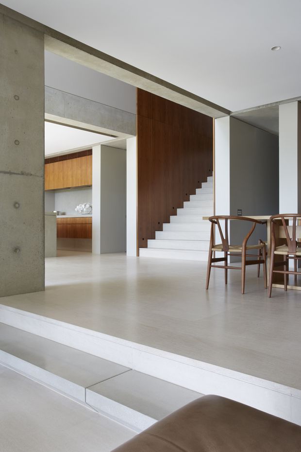 A bright, minimalist open-plan interior featuring a dining area in the foreground, a compact kitchen visible through a wide opening, and a white staircase set against a dark timber-panelled wall. Exposed concrete columns and beams frame the spaces, while pale flooring, white walls, and warm timber joinery create a restrained contemporary palette.
