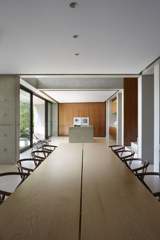 A minimalist open-plan interior viewed from a long dining table, with pale timber dining surfaces and dark sculptural chairs in the foreground. Exposed concrete walls and ceiling beams frame a contemporary kitchen beyond, featuring a central concrete island, extensive warm timber cabinetry, and large glazed openings bringing in greenery and daylight.