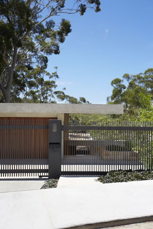 A contemporary residential front entrance viewed from the street, featuring a minimalist concrete slab canopy, vertical timber garage or entry screening, a dark metal pedestrian gate and sliding vehicle gate, and a sculptural dark mailbox or intercom pillar. Mature eucalyptus trees and landscaped planting surround the property, with strong sunlight and a clear blue sky.