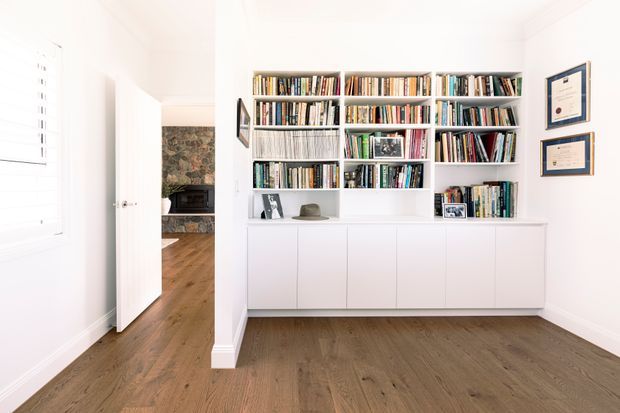 Bright, clean home office or study with a large built-in white bookshelf and lower storage cabinetry, filled with books and a few framed photographs. The room has white walls, framed certificates, timber flooring, and a doorway leading toward a stone-featured fireplace area.