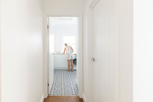 Bright white hallway viewed toward a laundry room, with patterned encaustic-style floor tiles, white cabinetry and a person standing at the laundry bench. A closed white paneled door and warm timber flooring are visible in the foreground.