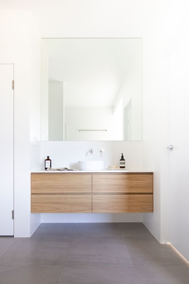 Bright minimalist bathroom featuring a wall-mounted natural timber vanity with integrated white basin, large frameless mirror, white tiled walls, and large grey floor tiles. The clean, uncluttered design includes wall-mounted tapware and subtle recessed lighting.