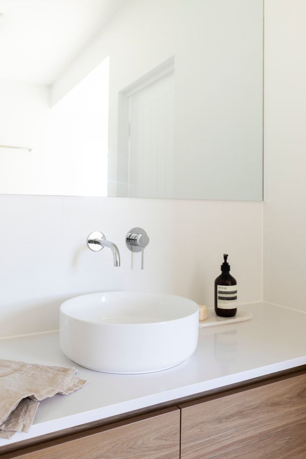 Bright, minimalist bathroom vanity with a white countertop, round white vessel basin, wall-mounted chrome tapware, large frameless mirror, and light timber cabinetry below. A dark amber soap dispenser and folded beige towel provide subtle contrast.