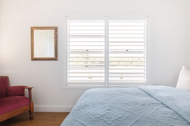 Bright, calm bedroom with white walls, a large white-framed plantation-shutter window, timber flooring, and a bed dressed in pale blue bedding. A traditional red upholstered armchair and decorative framed mirror add warm colour and character.