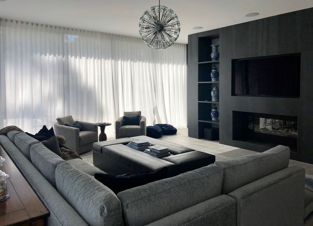 A sophisticated contemporary living room with a large grey sectional sofa arranged around a low dark coffee table. The room features full-height sheer curtains, a dark built-in media and fireplace wall with open display shelving, recessed ceiling lights, and a sculptural spherical pendant light. The palette is predominantly cool grey, charcoal, black, and white, creating a refined and calm atmosphere.