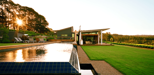 A landscaped rural property viewed from beside a long reflecting pool or swimming pool, with a low modern pavilion and separate dark-clad structure beyond. The building features broad roof overhangs, slender posts, extensive glazing, and a carefully maintained lawn and gravel pathway. Tall trees, planted garden beds, and rolling agricultural land form the setting, with warm low sunlight creating a serene evening atmosphere.